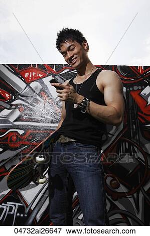 Young man standing in front of graffiti wall texting on phone while holding skateboard View Large Photo Image Stock Photo - Young man standing in front of graffiti wall texting on phone while holding skateboard. Fotosearch
