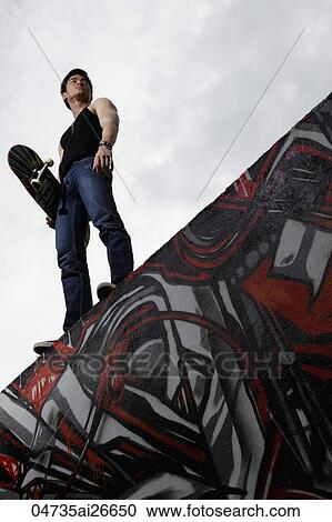 Young man standing on graffiti wall holding skateboard View Large Photo Image Stock Image - Young man standing on graffiti wall holding skateboard. Fotosearch