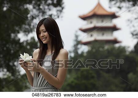 Stock Image - Young woman holding white flower in front of pagoda. Fotosearch