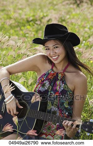 Stock Image - Smiling woman playing guitar in grassy field.. Fotosearch