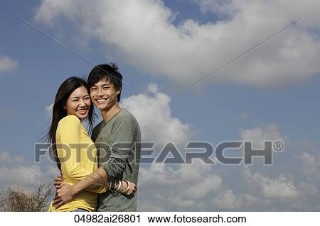 Stock Image - young couple hugging outside with blue sky and clouds as background. Fotosearch