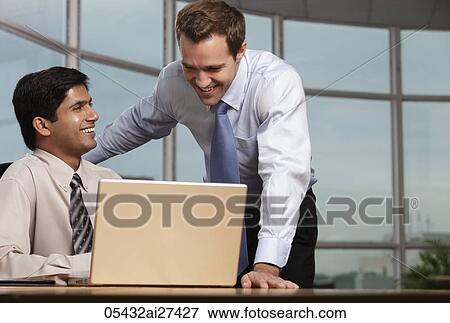 Stock Photo - Caucasian man looking at laptop with Indian man and smiling. Fotosearch