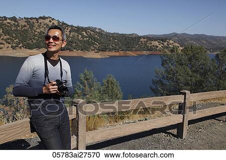 Stock Image - Chinese man standing near lake holding camera.. Fotosearch
