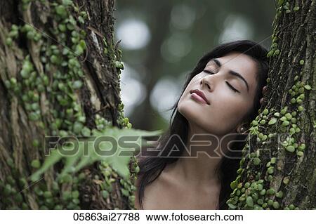Stock Photo - Head shot of young woman resting her head on a tree with eyes closed.. Fotosearch