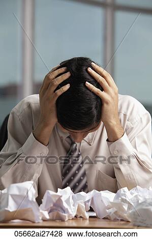 Stock Photo - Indian man holding head with crumpled up paper on table. Fotosearch