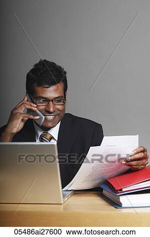 Stock Image - Indian man sitting at desk talking on phone and working on laptop. Fotosearch