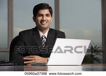 Stock Photo - Indian man sitting at table with laptop. Fotosearch