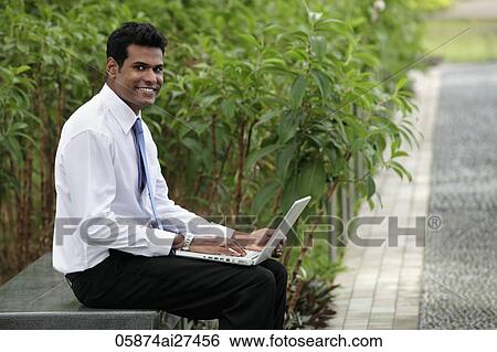 Stock Photograph - Indian man sitting on park bench working on laptop and smiling.. Fotosearch