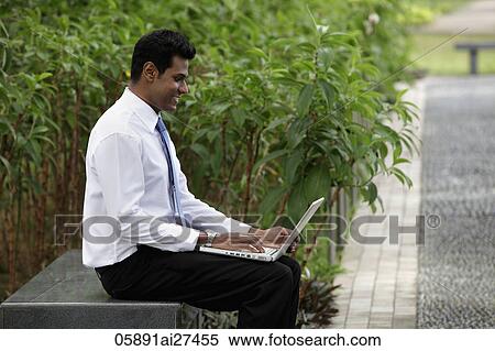 Stock Photography - Indian man sitting outside working on laptop with plants in background. Fotosearch