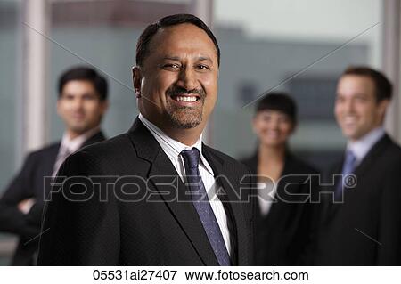 Stock Photo - Indian man smiling at camera with colleagues behind him. Fotosearch