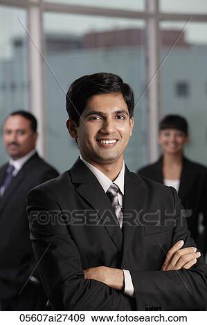 Stock Photo - Indian man smiling with arms folded. Colleagues behind him.. Fotosearch