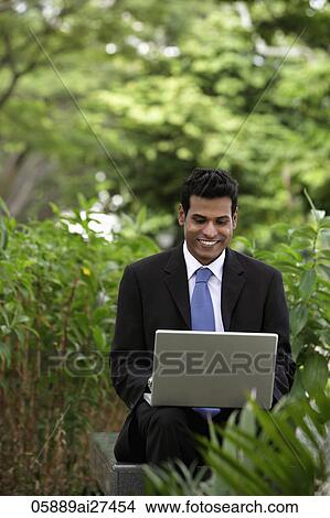 Indian man working on laptop with green plants and trees in foreground and background View Large Photo Image Picture - Indian man working on laptop with green plants and trees in foreground and background. Fotosearch