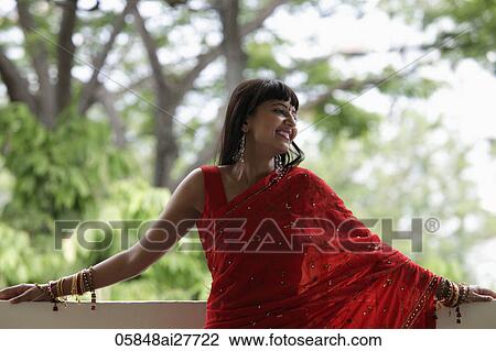 Stock Image - Indian woman wearing red sari leaning on balcony, smiling. Fotosearch