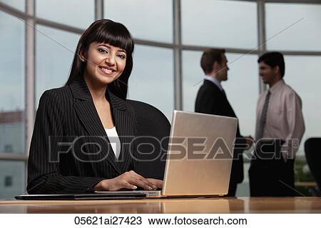 Stock Image - Indian woman working on laptop, colleagues in background. Fotosearch