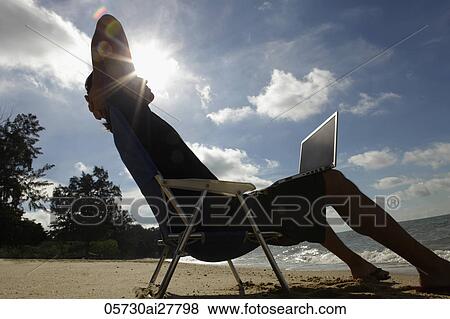 Stock Photo - Man sitting in beach chair with laptop, sun burst behind his head. Fotosearch