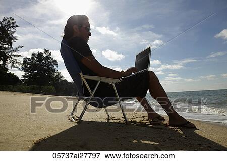 Man sitting on beach chair working on laptop at the beach View Large Photo Image Stock Photo - Man sitting on beach chair working on laptop at the beach. Fotosearch