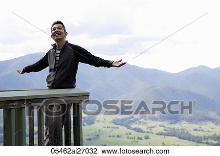 Stock Image - man smiling, standing on balcony with mountains as background. Fotosearch