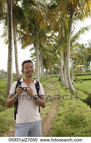 Stock Image - Man standing on a small path holding a camera in front of coconut trees.. Fotosearch