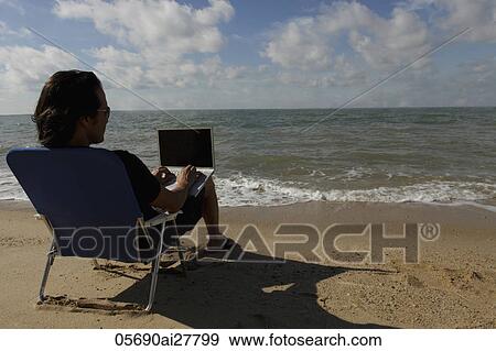 Rear view of man sitting on beach working on a laptop View Large Photo Image Stock Photo - Rear view of man sitting on beach working on a laptop. Fotosearch