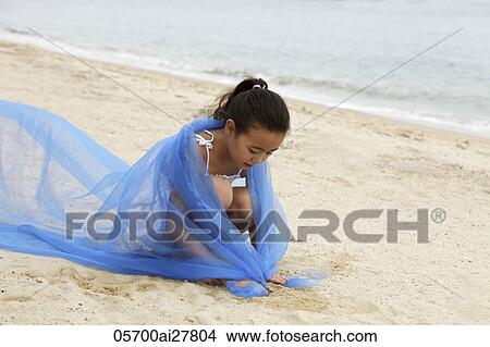 Picture - Young girl with blue cloth wrapped around her sitting on sand.. Fotosearch