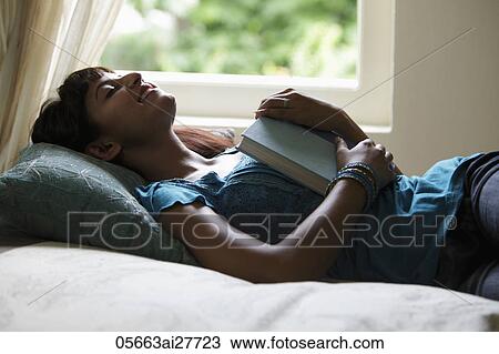 Stock Image - Young woman laying on back, smiling with eyes closed, holding book. Fotosearch