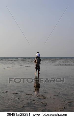 Stock Image - Back vier of boy sitting on dad's shoulders looking at the ocean. Fotosearch