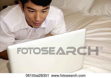 Stock Image - man laying on bed working on laptop. Fotosearch