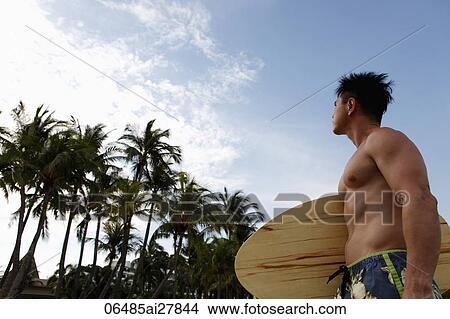 man wearing swim suit holding skim board looking at palm trees View Large Photo Image Picture - man wearing swim suit holding skim board looking at palm trees. Fotosearch