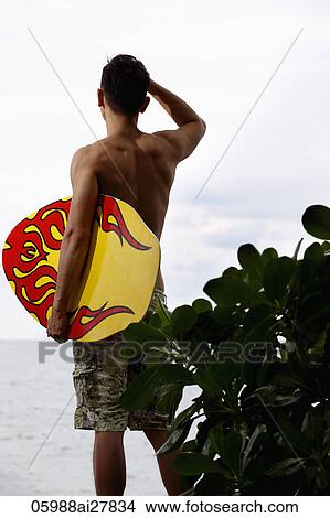 rear view of young man holding skim board looking at ocean View Large Photo Image Picture - rear view of young man holding skim board looking at ocean. Fotosearch