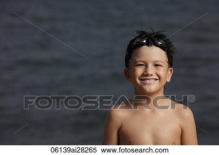 Stock Photography - Young boy standing in front of ocean smiling with goggles on his head. Fotosearch