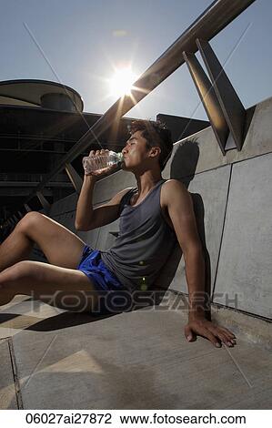Stock Image - Young man leaning against wall and drinking water. Fotosearch