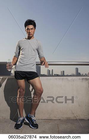 Picture - Young man leaning against wall holding water bottle. Fotosearch