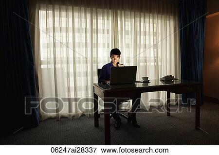 Young man sitting at desk working on laptop in the dark View Large Photo Image Stock Photo - Young man sitting at desk working on laptop in the dark. Fotosearch