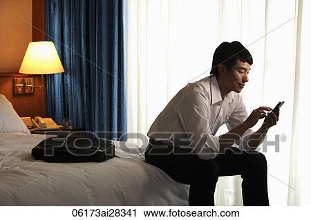 Stock Image - young man sitting on bed in hotel room texting on phone. Fotosearch