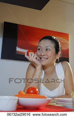 Stock Photograph - Chinese woman sitting at table holding tomato and smiling. Fotosearch