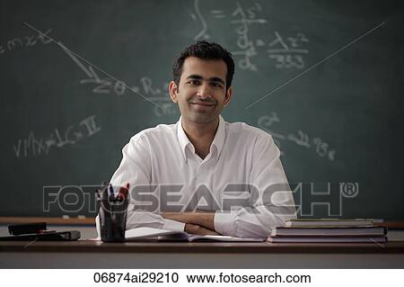 Stock Image - Young man sitting at his desk smiling with chalk board behind. Fotosearch