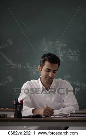 Young man sitting at his desk writing with chalk board in background. View Large Photo Image Stock Image - Young man sitting at his desk writing with chalk board in background.. Fotosearch