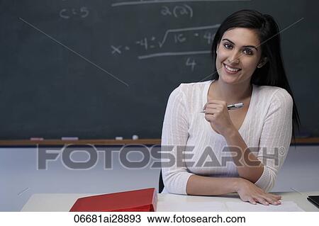 Stock Image - Young teacher sitting at desk in front of chalkboard and smiling. Fotosearch