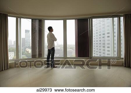 Stock Image - Rear view of young man looking out large windows of condo. Fotosearch