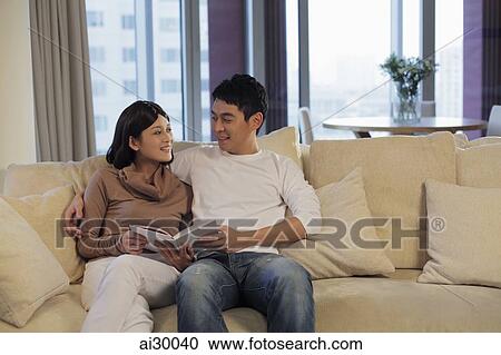 Stock Image - Young couple reading a book together on a sofa. Fotosearch
