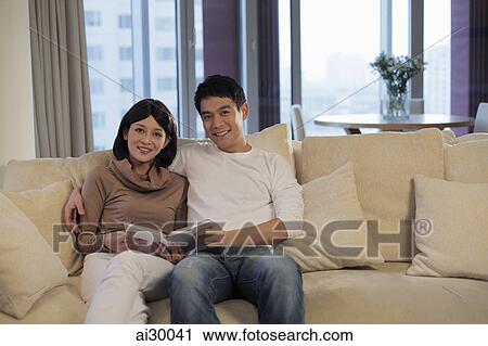 Stock Image - Young couple sitting on sofa reading a book. Fotosearch