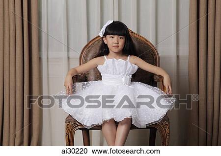 Stock Image - Young girl dressed up in white dress and sitting on nice chair. Fotosearch