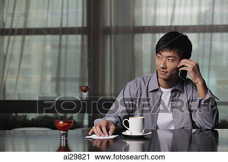 Stock Image - Young man sitting at a table talking on the phone. Fotosearch