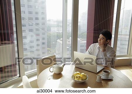 Young man sitting at a table with a computer looking out the window View Large Photo Image Picture - Young man sitting at a table with a computer looking out the window. Fotosearch