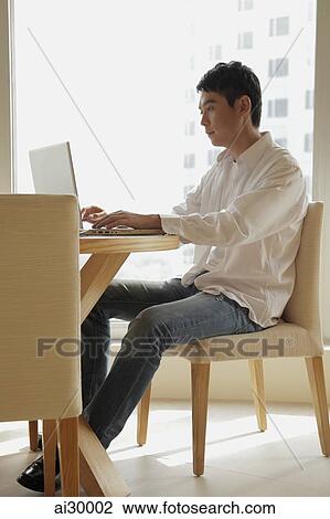Young man sitting at a table working on a laptop computer View Large Photo Image Stock Image - Young man sitting at a table working on a laptop computer. Fotosearch