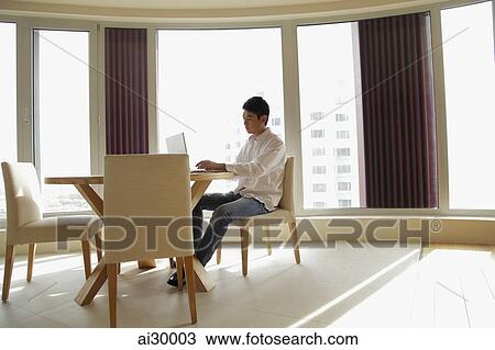Young man sitting at a table working on a laptop computer View Large Photo Image Stock Image - Young man sitting at a table working on a laptop computer. Fotosearch
