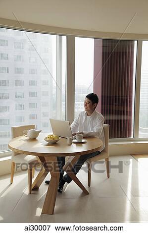 Young man sitting at a table working on a laptop computer View Large Photo Image Stock Photo - Young man sitting at a table working on a laptop computer. Fotosearch