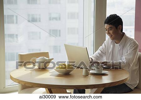 Young man sitting at a table working on a laptop computer View Large Photo Image Stock Photo - Young man sitting at a table working on a laptop computer. Fotosearch