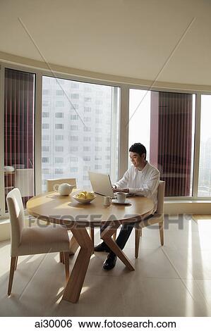Young man sitting at a table working on a laptop computer View Large Photo Image Stock Photograph - Young man sitting at a table working on a laptop computer. Fotosearch