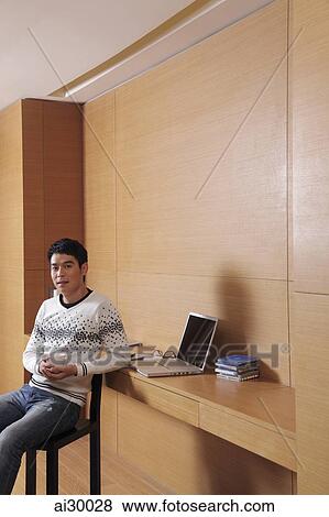 Young man sitting down at desk holding coffee View Large Photo Image Stock Photo - Young man sitting down at desk holding coffee. Fotosearch